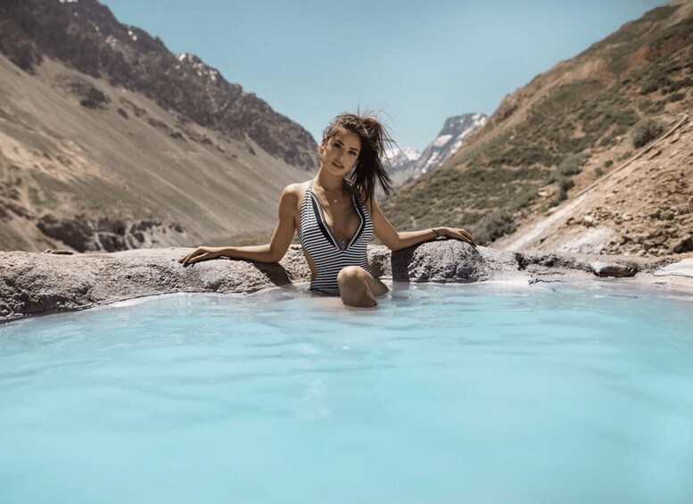 A person sitting in a clear blue pool surrounded by rocky terrain and mountains. 1