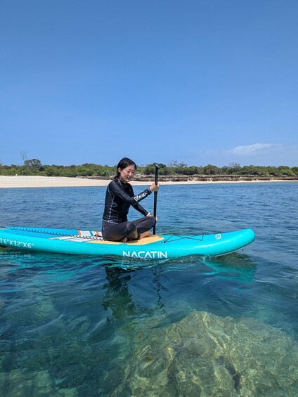 A person sitting on a paddleboard in clear water with a beach and trees in the background. 1