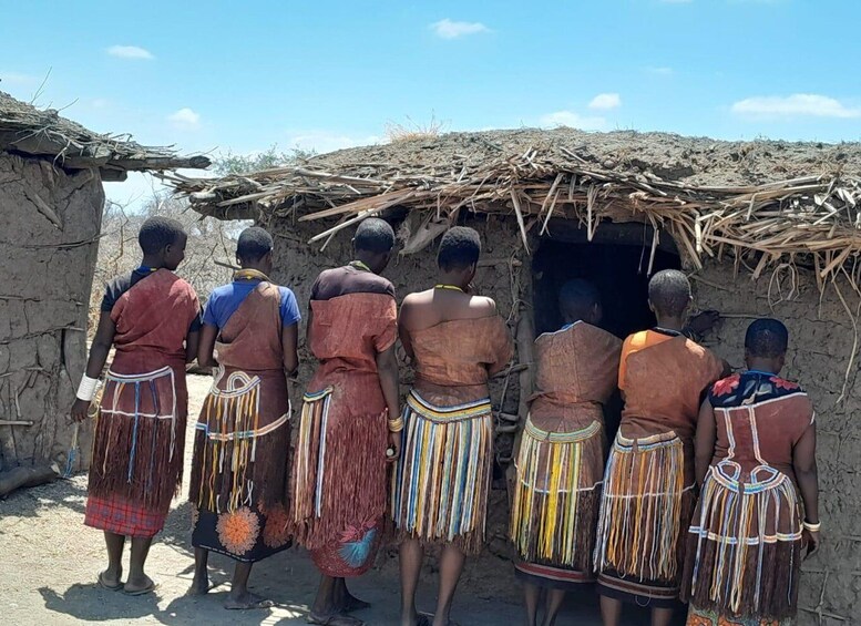 A group of people standing in front of a traditional mud hut with a thatched roof. 2