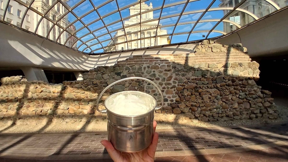 A hand holding a metal cup in front of a stone wall with a glass ceiling structure in the background. 1
