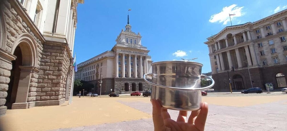 A hand holding a metallic cup in front of a building with columns and a spire. 1