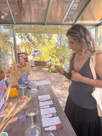 A person standing at a table with wine glasses and informational pamphlets, outdoors with trees and a bench in the background. 2