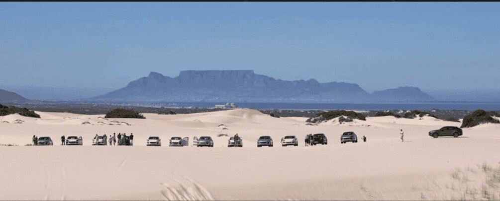A desert landscape with a line of cars parked on the sand, a mountain range in the background, and sparse vegetation. 1