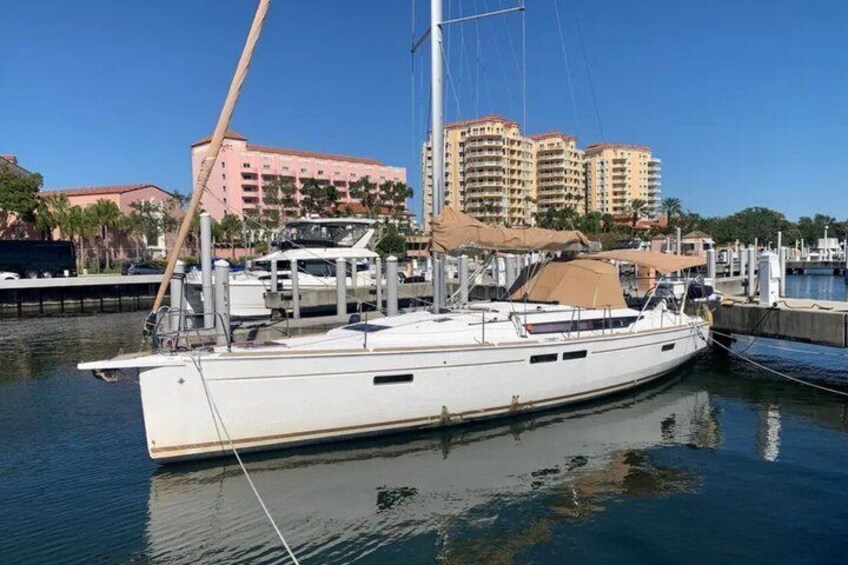 Een witte zeilboot afgemeerd in een jachthaven, met gebouwen op de achtergrond. 1