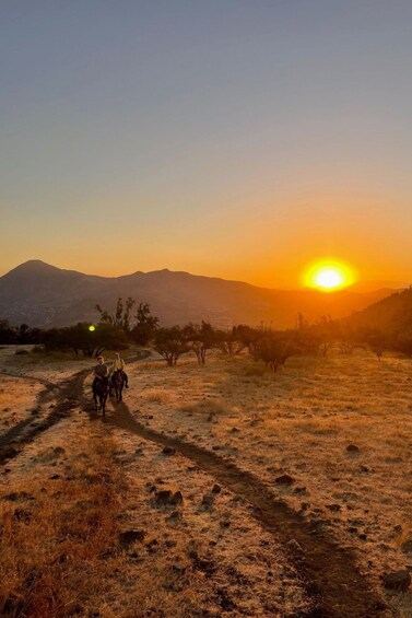 A sunset over a mountainous landscape with a dirt path and two riders on horseback. 1