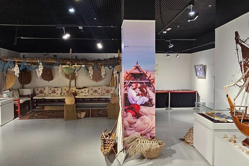 Eine Innenansicht einer Ausstellung mit traditionellen Artefakten, darunter ein großes Gemälde, gewebte Körbe und ein Modellschiff. 2