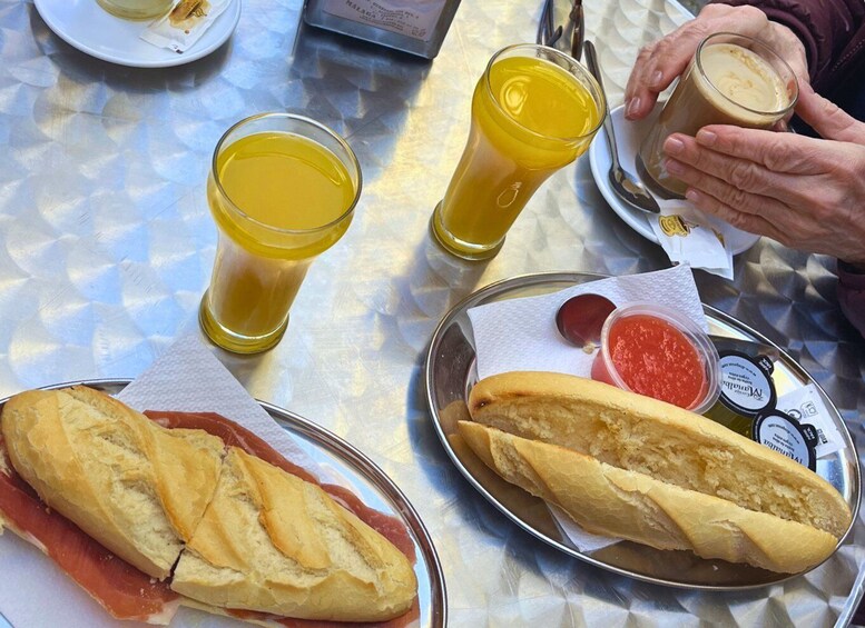 A table with food and drinks. There are two baguettes, one with smoked salmon and the other with a red sauce. There are also two glasses of orange juice and a cup of coffee. 1