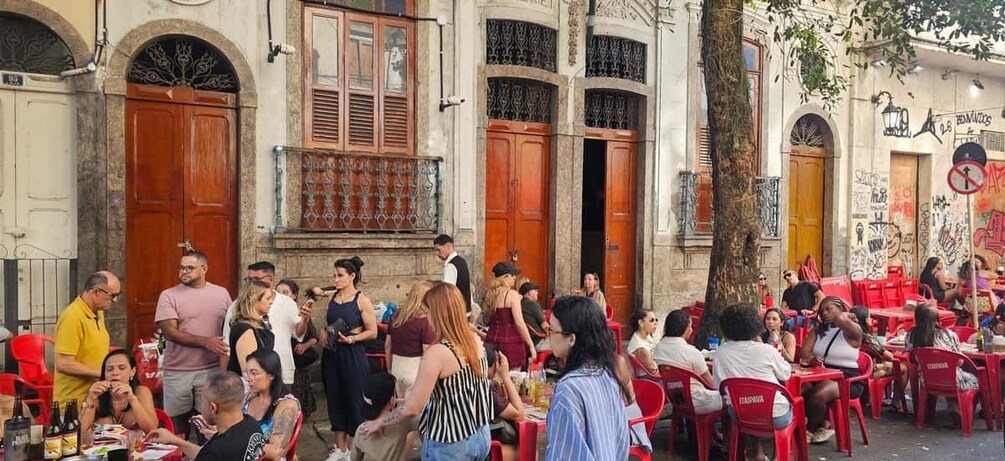 A crowded outdoor cafe with red chairs and tables, surrounded by buildings with large wooden doors and windows. 4