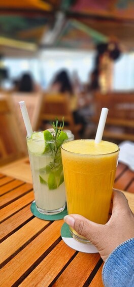 A person holding two drinks on a wooden table with a blurred background of people. 2