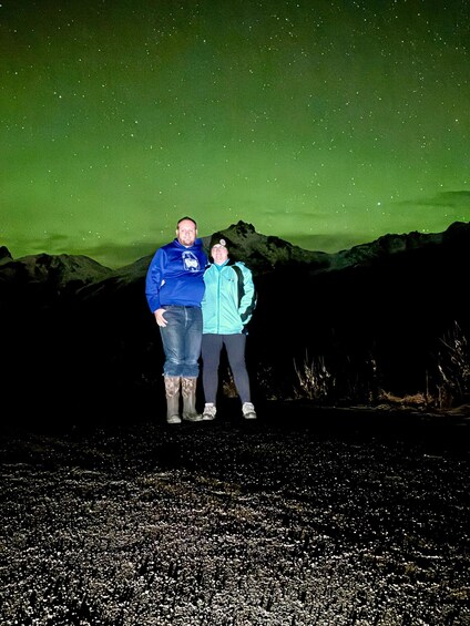Two people standing on a rocky terrain under a starry night sky with a green aurora. 2
