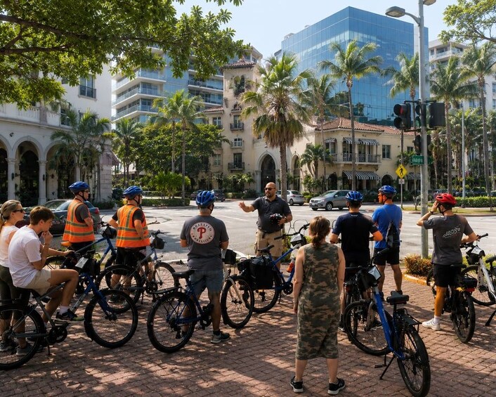 A group of cyclists gathered on a city street with palm trees and buildings in the background. 5