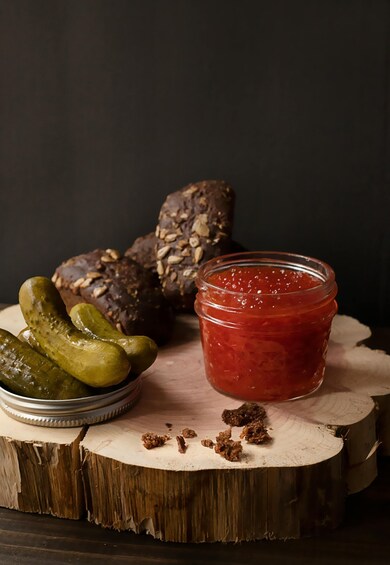 A jar of red jam, pickles, and a loaf of bread on a wooden surface. 1