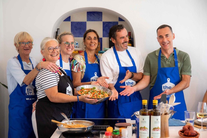 A group of people wearing aprons with 'the' logo, standing around a table with food and cooking utensils. 1