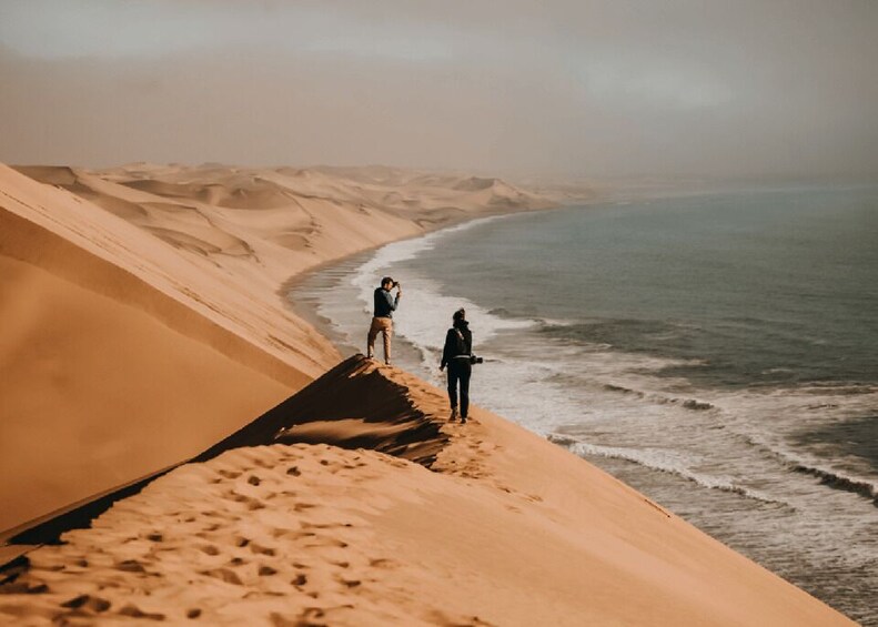 Two people standing on a sand dune overlooking a beach with waves. 1