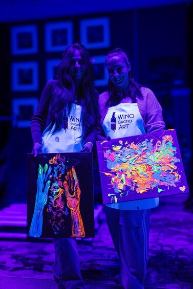 Two people holding colorful paintings in a dimly lit room with purple lighting. 2
