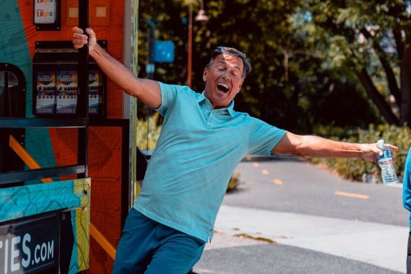 Un hombre con una camisa azul apoyado contra un autobús colorido, sosteniendo una botella de agua. 3