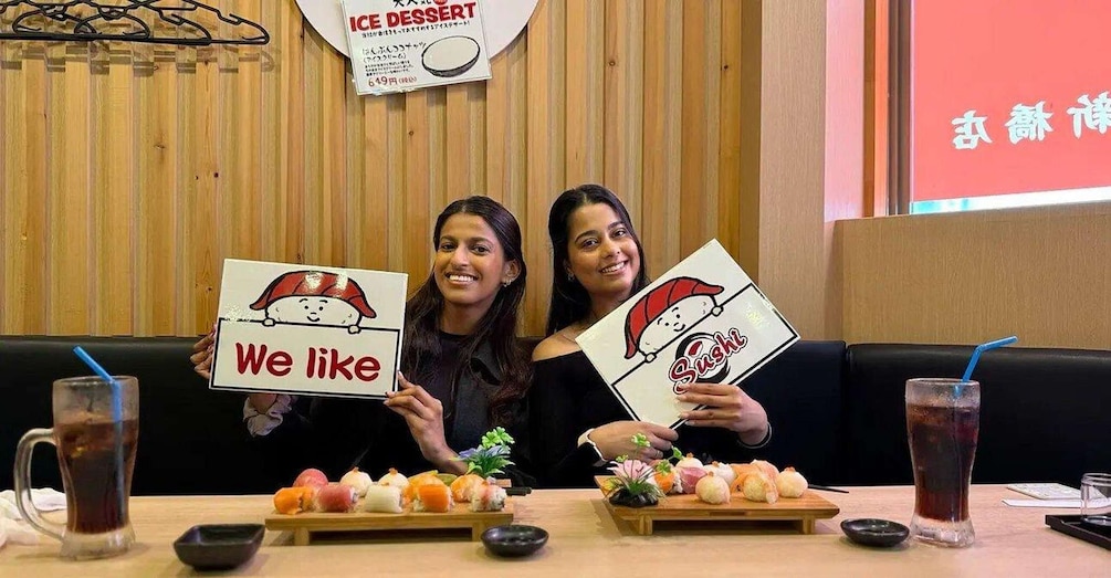 Two people holding signs with food illustrations, a sushi platter, and a menu board in a restaurant. 4