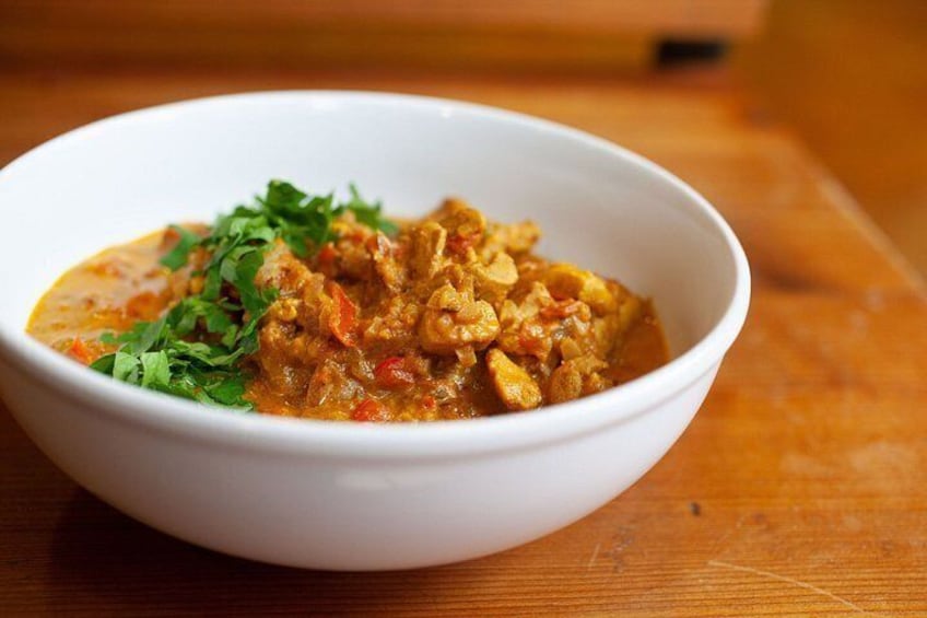 A white bowl of curry with vegetables and herbs on a wooden table. 1
