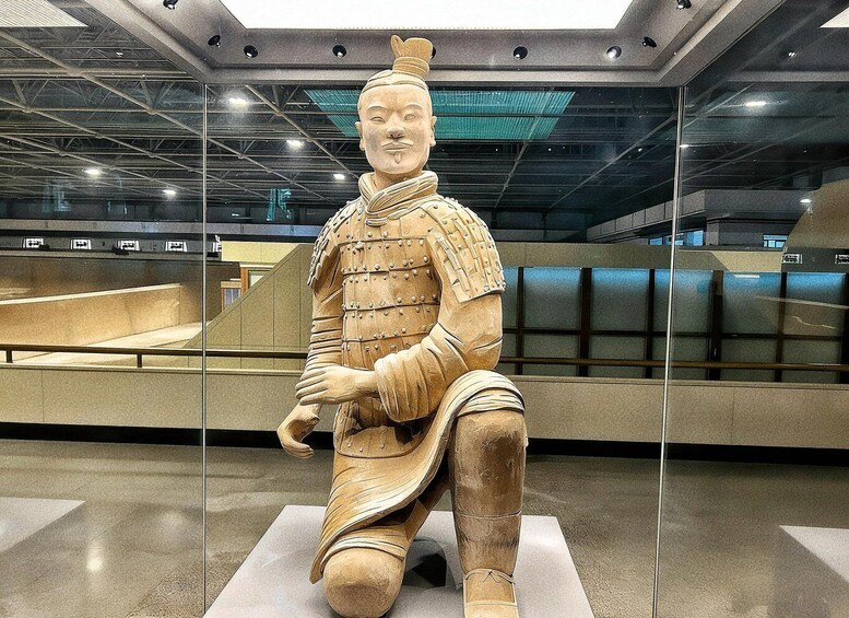 A terracotta statue of a seated figure in armor, displayed indoors with a modern ceiling and glass walls. 1