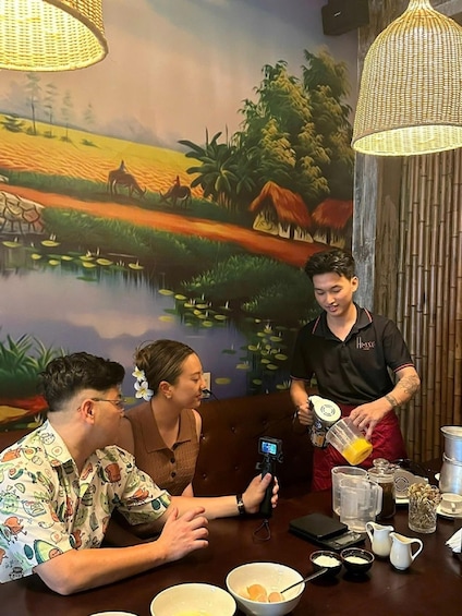 A person pouring a drink into a glass at a table with a scenic mural in the background. 1