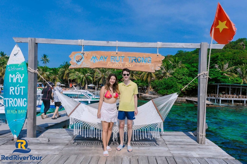 A wooden dock with a hammock, palm trees, and a sign for May Rut Trong Beach Bar. 2