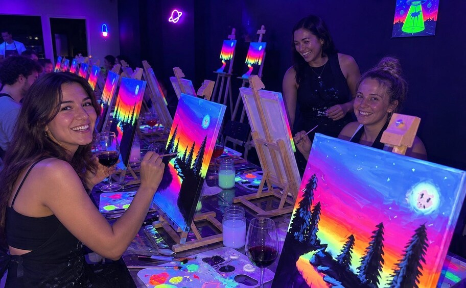 People painting at a table with easels and canvases, in a dimly lit room with neon signs. 1