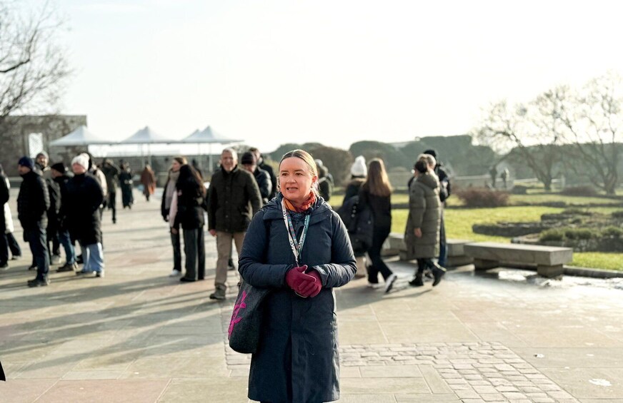 A person in a blue coat with a pink bag standing on a paved area with people walking in the background. 3