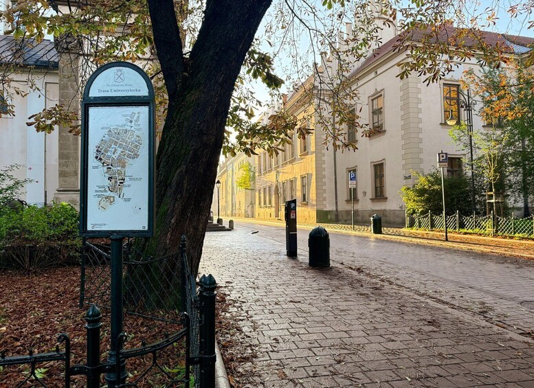 A tree-lined cobblestone street with a map sign, buildings, and a parking sign. 1