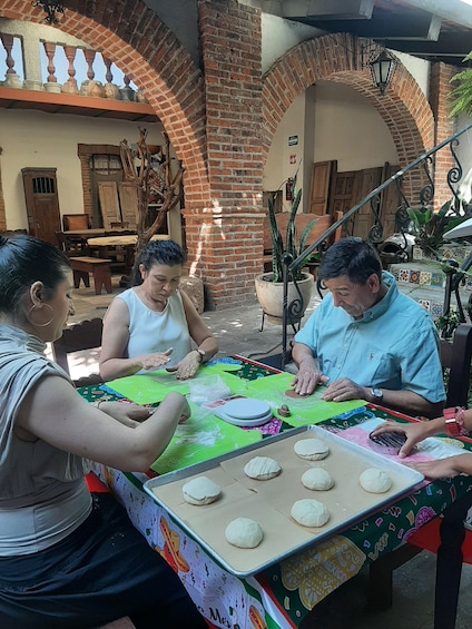 A group of people engaged in a dough preparation activity at a table with a colorful tablecloth, surrounded by brick arches and potted plants. 1