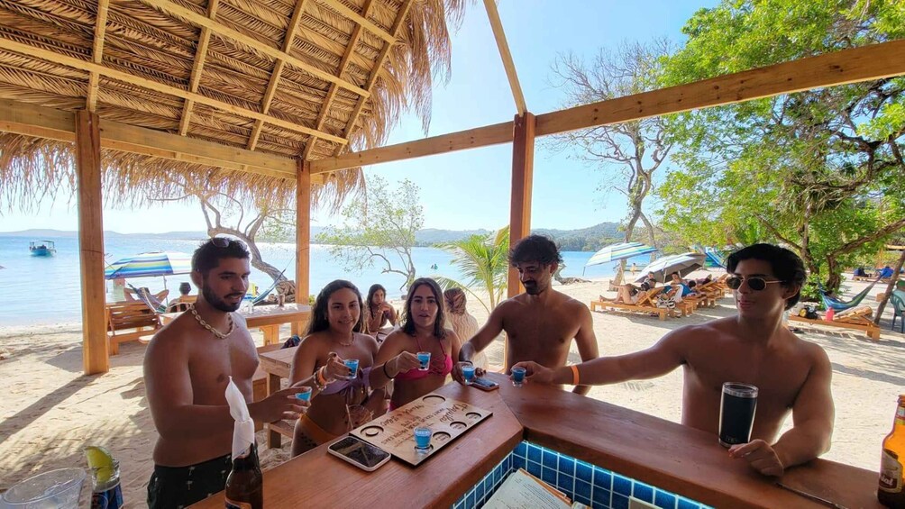 A group of people at a beach bar with a thatched roof, wooden furniture, and a view of the sea. 5