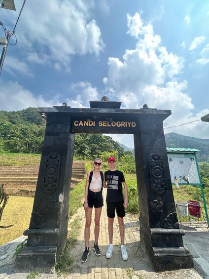 Two people standing under a stone archway with 'Candi Selo Griyo' inscribed on it, surrounded by terraced fields and a clear sky. 2