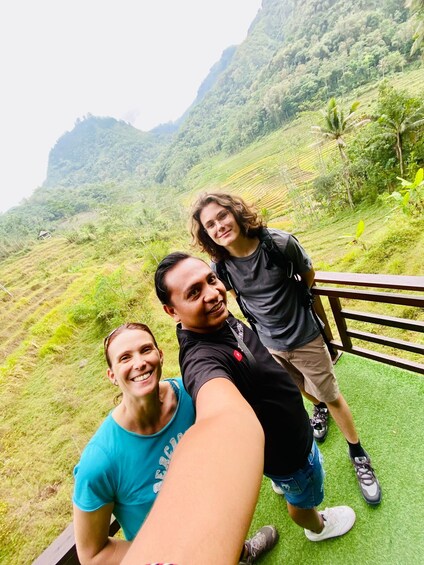 Three people taking a selfie on a wooden platform with a lush green landscape and mountains in the background. 4