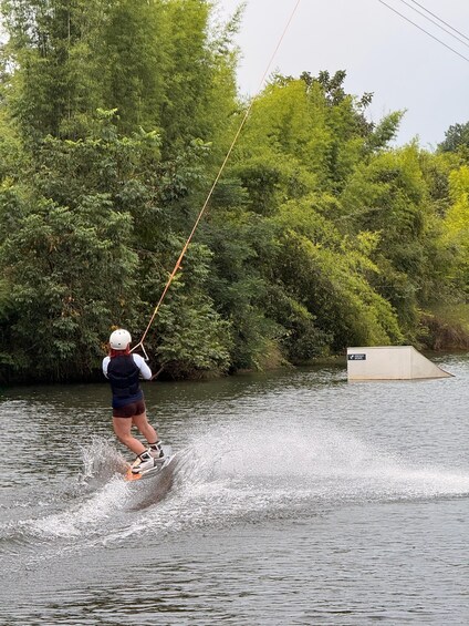 A person wakeboarding on a river, wearing a helmet and life jacket, with trees and a ramp in the background. 1
