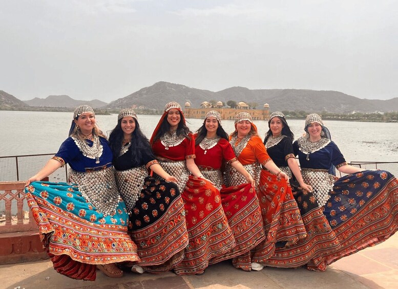 A group of women in traditional attire by a lakeside with a palace in the background. 3
