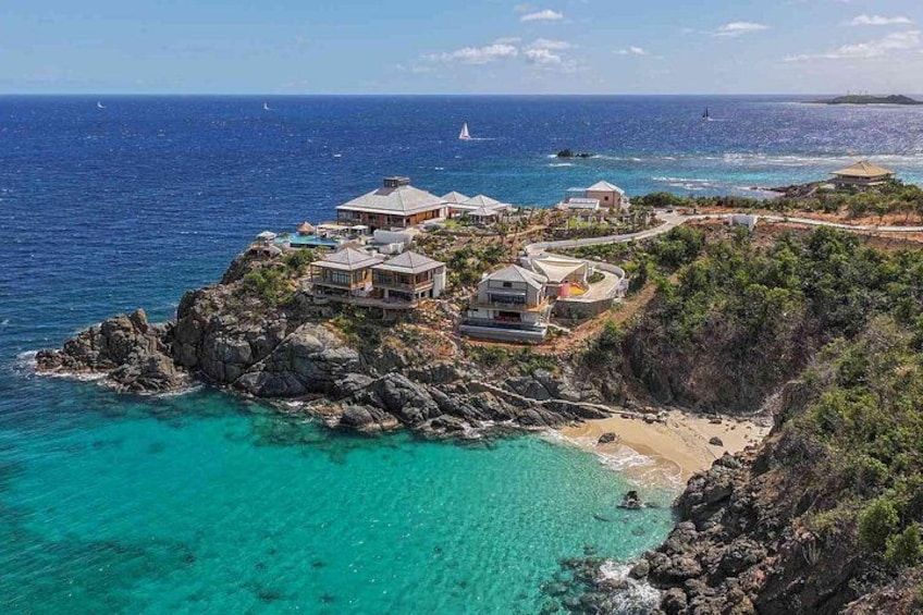 Un complejo turístico frente al mar con edificios sobre un promontorio rocoso, aguas turquesas cristalinas y una playa de arena. 2