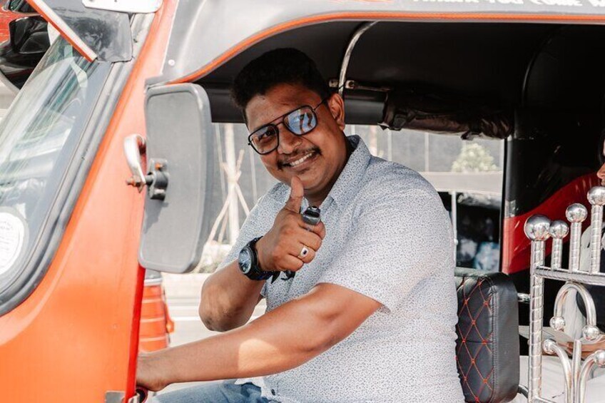 Een man zittend in een oranje tuktuk, die een duimpje omhoog steekt. 1