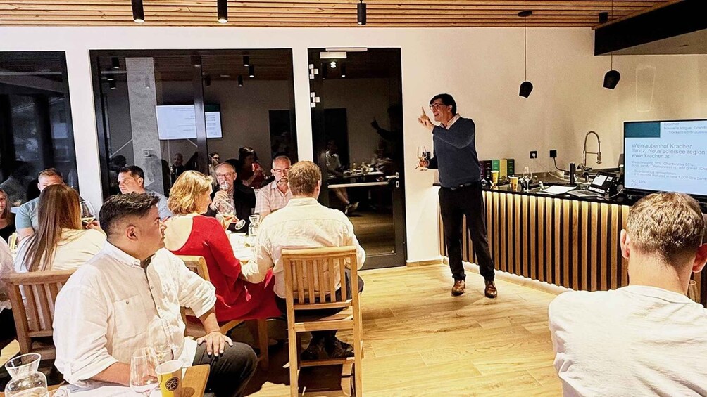 A person presenting in a modern conference room with wooden floors and tables, attendees seated, and a digital display on the wall. 2