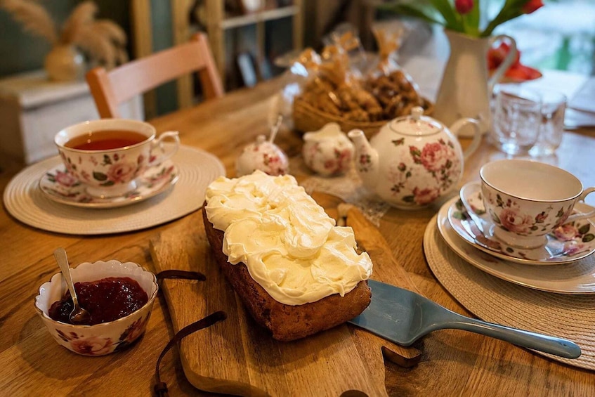 A wooden table with a slice of bread topped with cream cheese, a teapot, cups, and a bowl of jam. 2