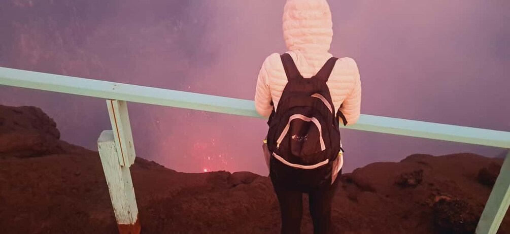 A person in a hooded jacket and backpack standing at a railing, overlooking a reddish landscape. 3
