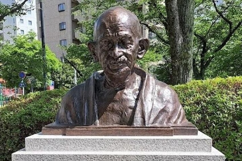 Une statue de bronze représentant un homme, placée sur un piédestal dans un parc, avec des arbres et des bâtiments en arrière-plan. 2