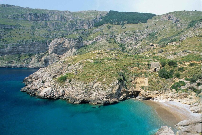 A coastal landscape with a rocky shoreline, clear blue waters, and a small building on a hill. 4