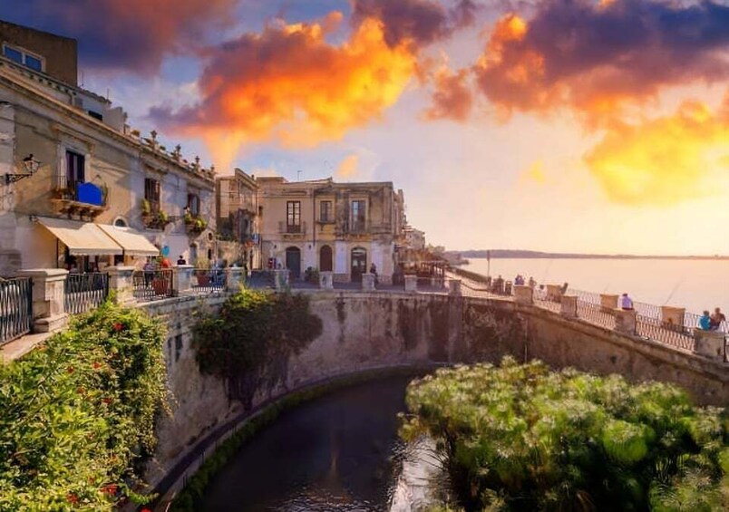A waterfront promenade with buildings, a curved canal, and a sunset sky. 1