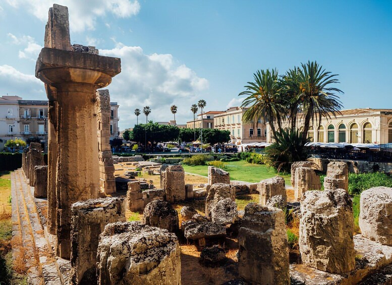 Ancient ruins with stone columns and a large stone structure, surrounded by palm trees and modern buildings. 5