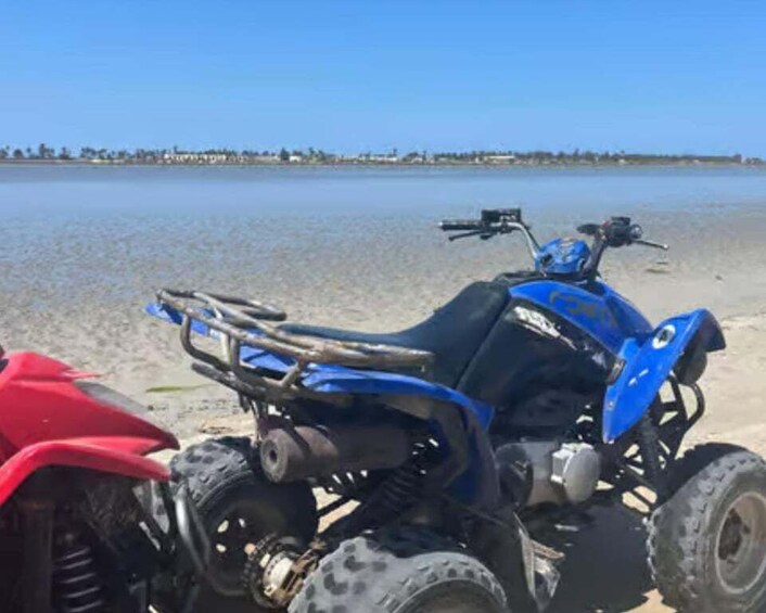 Un vehículo todo terreno azul estacionado en una playa arenosa, con una masa de agua y una costa distante al fondo. 2