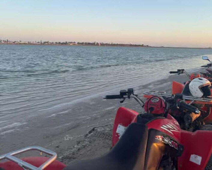 Un vehículo todo terreno rojo estacionado en una playa arenosa con agua tranquila al fondo. 3