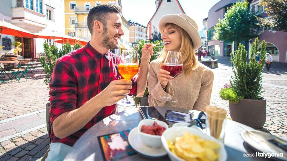A couple sitting at an outdoor café, holding glasses of wine, with a table full of food and a city street in the background. 4