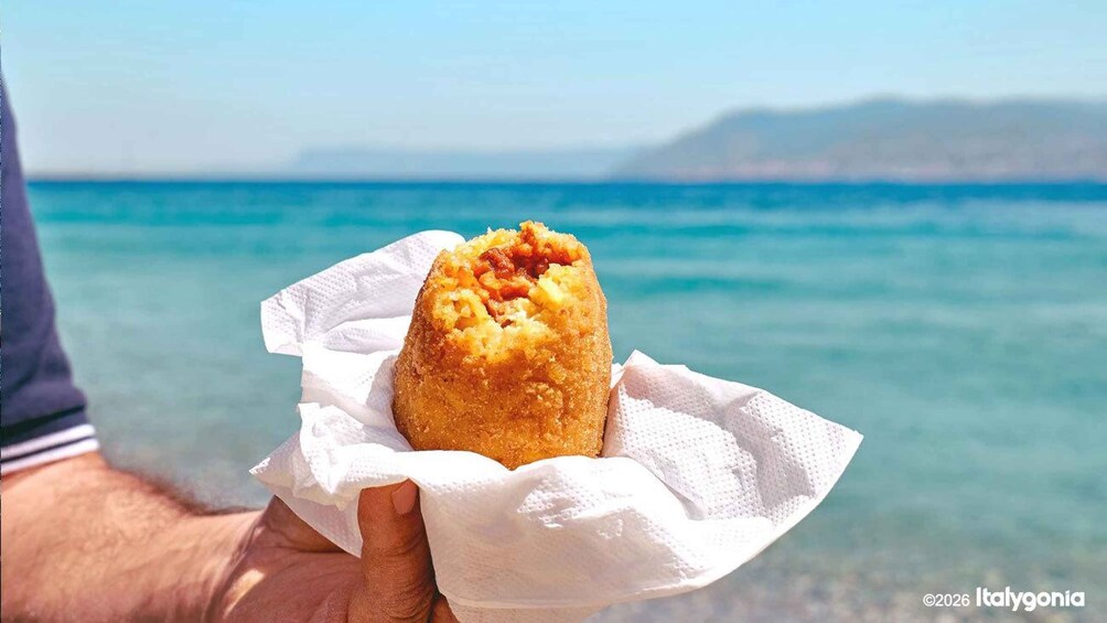 A person holding a fried food item with a clear blue sea and distant mountains in the background. 1