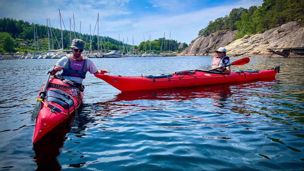Two people kayaking on a calm lake with boats and a rocky shoreline in the background. 2