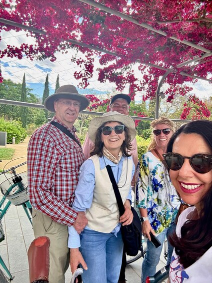 A group of people standing under a pergola with pink flowers, with a bicycle and greenery in the background. 2