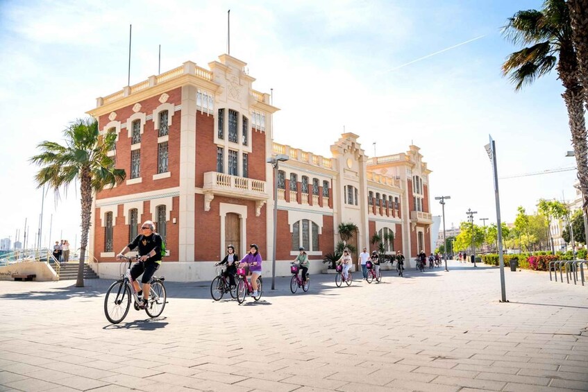 A group of people riding bicycles on a paved walkway in front of a large, ornate building with a red and beige facade. 5